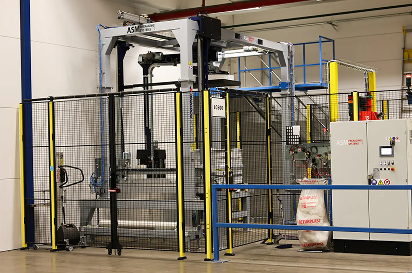 Automatiserat förpackningssystem i ett lager, inhägnat med säkerhetsstängsel och utrustat med kontrollpaneler.